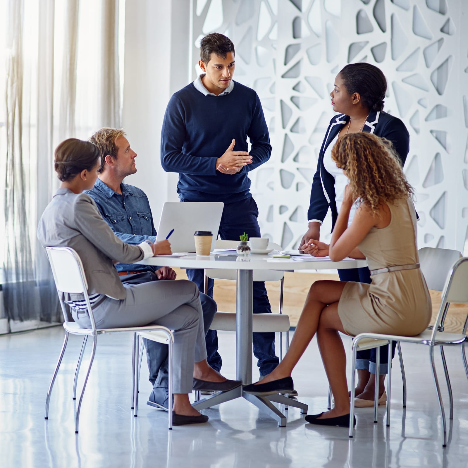 The image shows a bright, modern conference room with five diverse professionals in discussion on hiring strategy. They are seated around a white table, with papers and laptops present, conveying a serious yet optimistic atmosphere of partnering with HireCanny. Natural light enters through large windows, complemented by soft overhead lighting and modern art on the walls. The professionals are dressed impeccably, captured in high resolution and sharp focus, highlighting their focused expressions.